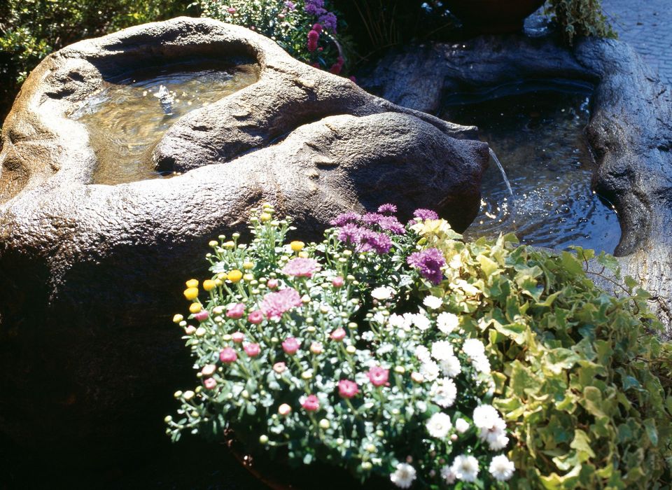 Stone water fountain with flowing water surrounded by colorful flowers and green plants Stone water fountain with flowing water surrounded by colorful flowers and green plants