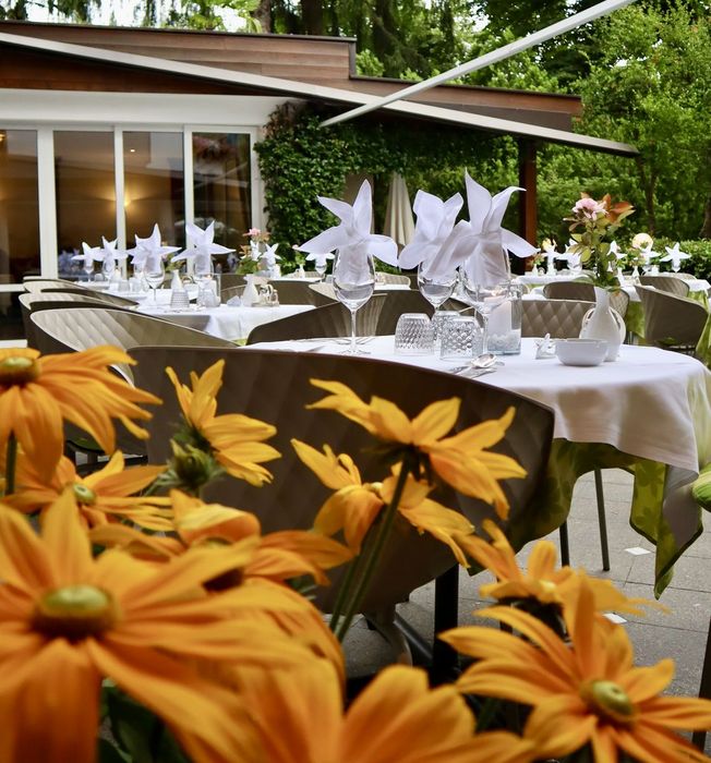 Outdoor restaurant seating with white tablecloths and floral centerpieces surrounded by green trees and yellow flowers in the foreground. Outdoor restaurant seating with white tablecloths and floral centerpieces surrounded by green trees and yellow flowers in the foreground.