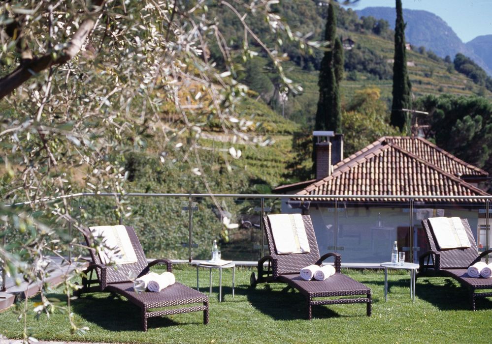 Outdoor patio with three lounge chairs on grass, towels, and small tables, overlooking a scenic hillside with trees and houses. Outdoor patio with three lounge chairs on grass, towels, and small tables, overlooking a scenic hillside with trees and houses.