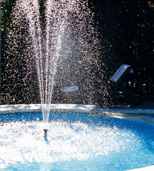 Water fountain spraying water droplets in a pool with greenery in the background. Water fountain spraying water droplets in a pool with greenery in the background.